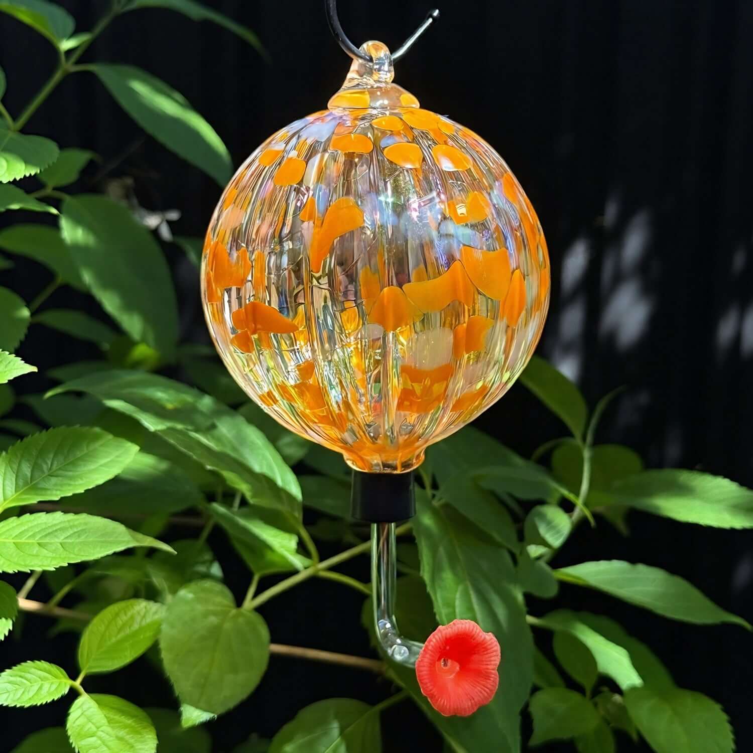 Handmade Salmonberry glass art from the Lacey Collection, featuring vibrant colors hanging in garden foliage.